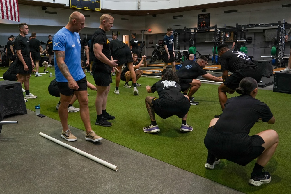 A group of military personnel are participating in a structured physical training session inside a modern gymnasium facility. Instructors in blue shirts appear to be supervising and coaching soldiers dressed in black PT uniforms performing squat and mobility exercises. The setting appears to be a military fitness center, evidenced by Army insignia on clothing and an American flag visible in the background.