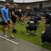 A group of military personnel are participating in a structured physical training session inside a modern gymnasium facility. Instructors in blue shirts appear to be supervising and coaching soldiers dressed in black PT uniforms performing squat and mobility exercises. The setting appears to be a military fitness center, evidenced by Army insignia on clothing and an American flag visible in the background.