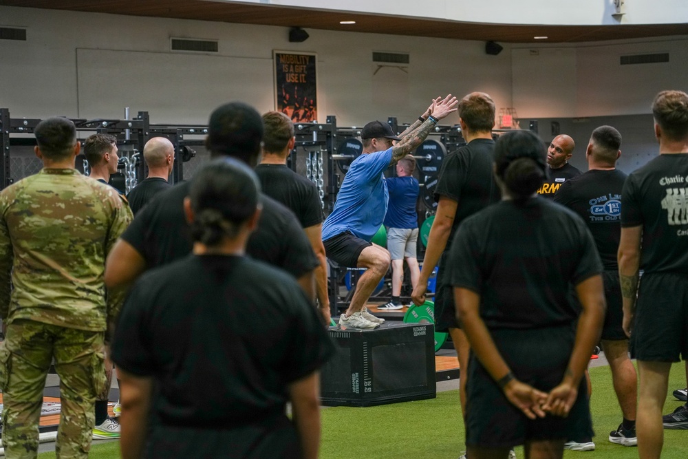A fitness instructor in a blue shirt is demonstrating a box jump exercise on a plyo box inside a modern military gym facility. A group of military personnel and civilians in black athletic wear and one person in camouflage uniform observe the demonstration. The setting appears to be a formal physical training or fitness education session.