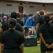 A fitness instructor in a blue shirt is demonstrating a box jump exercise on a plyo box inside a modern military gym facility. A group of military personnel and civilians in black athletic wear and one person in camouflage uniform observe the demonstration. The setting appears to be a formal physical training or fitness education session.