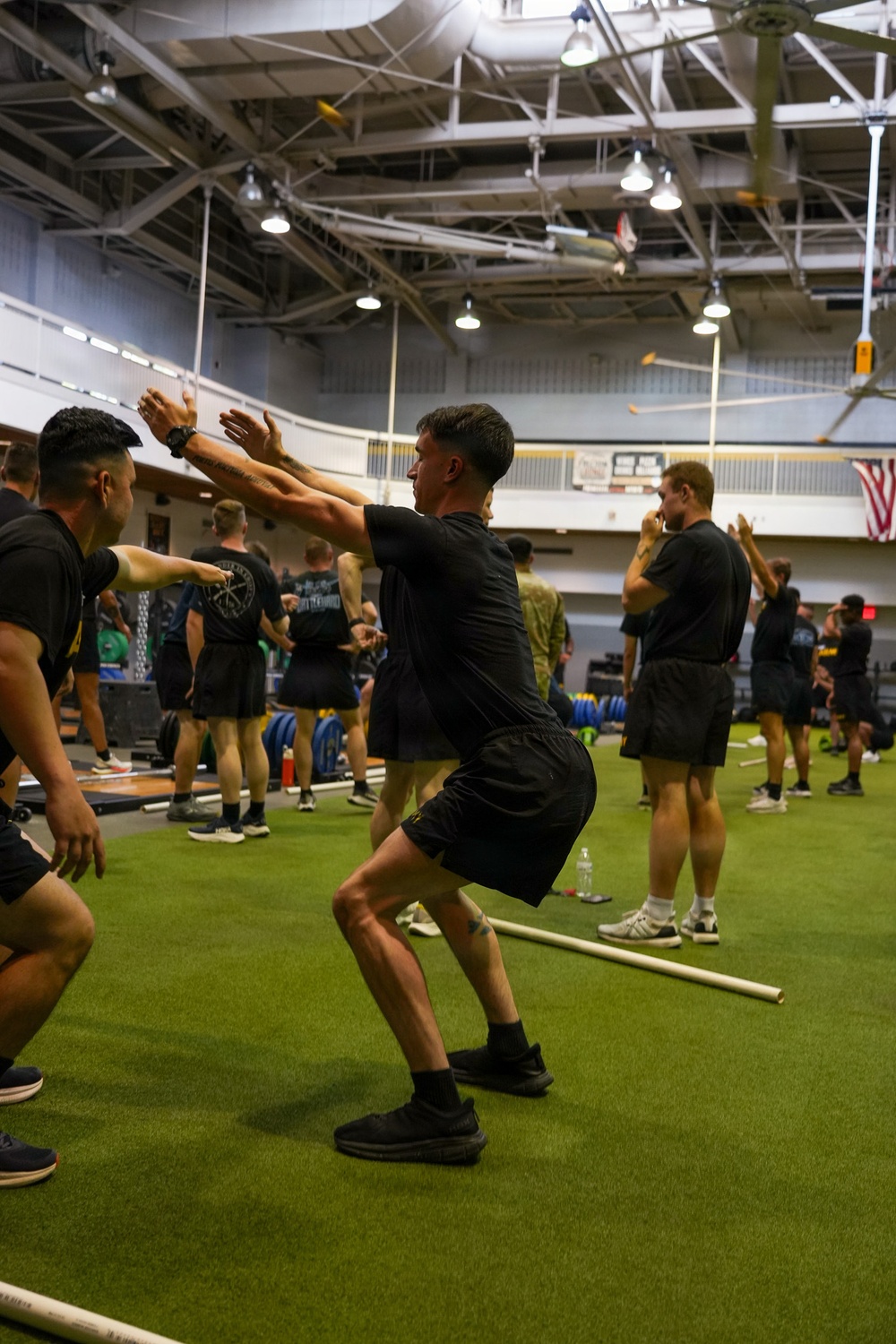 A large group of individuals dressed in matching black athletic uniforms are participating in a group physical training session inside a large indoor gymnasium facility. The participants appear to be performing squat or movement exercises in an organized group format on an artificial turf surface. An American flag is visible in the background, suggesting this may be a military or government-affiliated fitness facility.