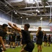 A large group of individuals dressed in matching black athletic uniforms are participating in a group physical training session inside a large indoor gymnasium facility. The participants appear to be performing squat or movement exercises in an organized group format on an artificial turf surface. An American flag is visible in the background, suggesting this may be a military or government-affiliated fitness facility.