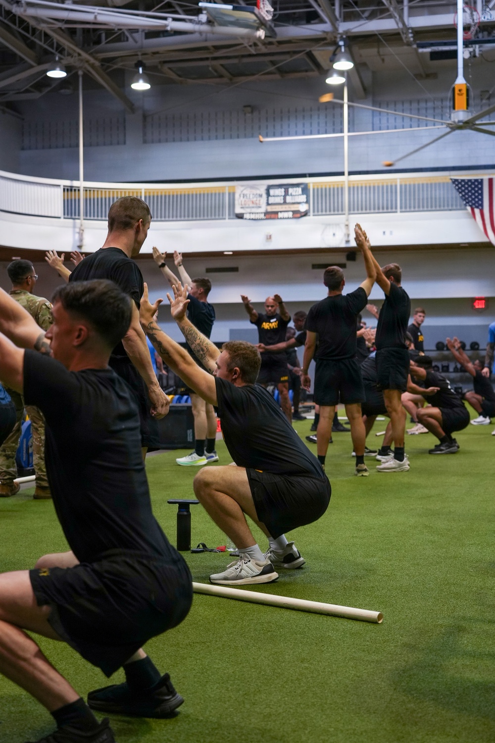 A large group of military personnel in black Army PT (Physical Training) uniforms are performing synchronized squat exercises inside an indoor sports facility. The participants are in various stages of a squat movement with arms raised, suggesting a structured group physical training session. An American flag and an 'ARMY' branded shirt are visible, indicating a U.S. Army training environment.