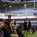 A large group of military personnel in black Army PT (Physical Training) uniforms are performing synchronized squat exercises inside an indoor sports facility. The participants are in various stages of a squat movement with arms raised, suggesting a structured group physical training session. An American flag and an 'ARMY' branded shirt are visible, indicating a U.S. Army training environment.