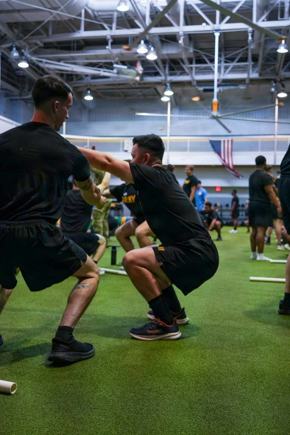 A group of individuals wearing black U.S. Army physical training uniforms are engaged in a partner-assisted squat or resistance exercise inside a large indoor gymnasium facility. The facility features artificial turf flooring, high ceilings with industrial lighting, and an American flag visible in the background. Multiple pairs of participants are performing the same exercise simultaneously, suggesting a structured group physical training session.