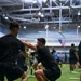 A group of individuals wearing black U.S. Army physical training uniforms are engaged in a partner-assisted squat or resistance exercise inside a large indoor gymnasium facility. The facility features artificial turf flooring, high ceilings with industrial lighting, and an American flag visible in the background. Multiple pairs of participants are performing the same exercise simultaneously, suggesting a structured group physical training session.
