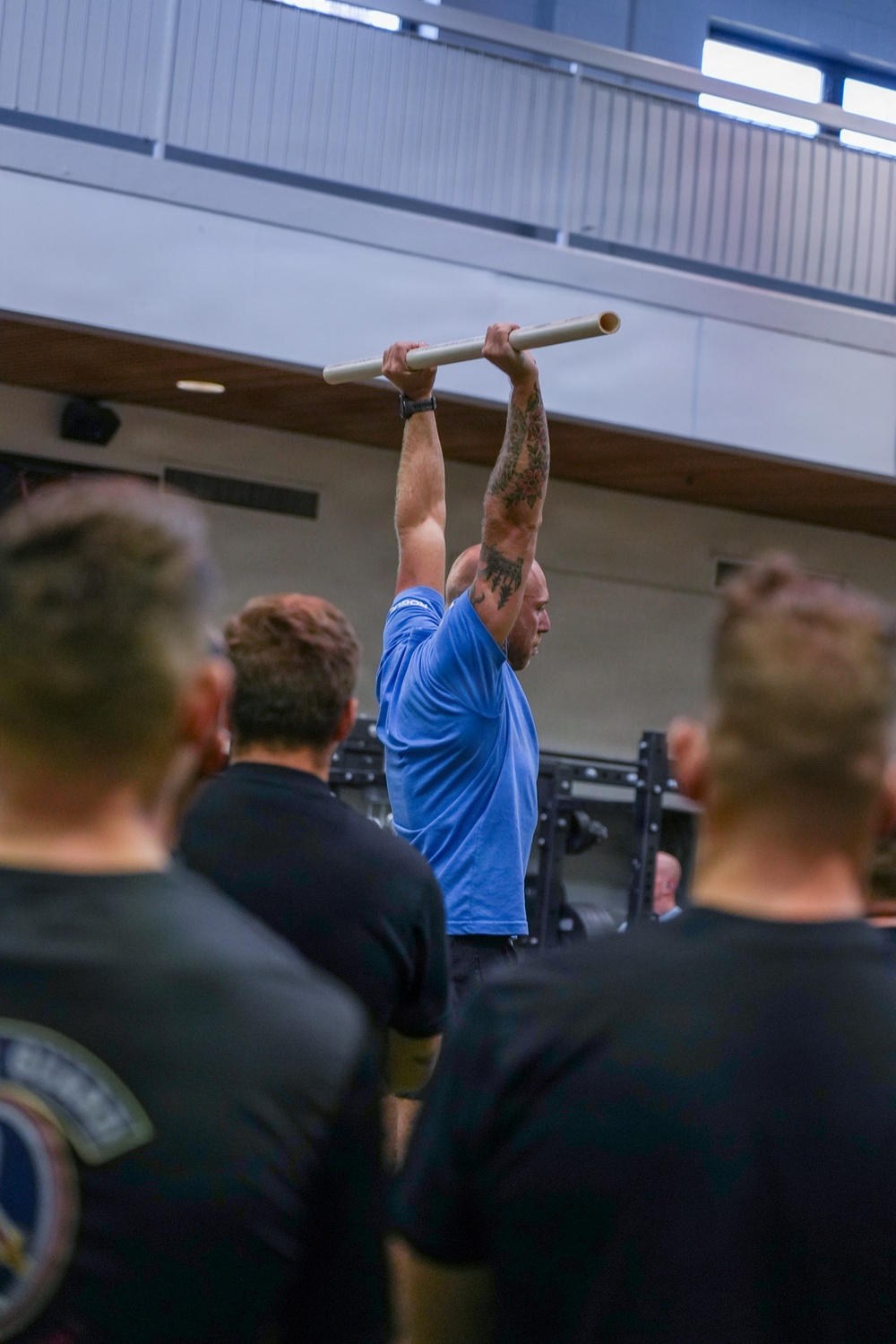 A muscular man wearing a blue t-shirt is performing a dead hang or pull-up on a horizontal bar in what appears to be an indoor gymnasium or fitness facility. He has visible tattoos on his right arm and is wearing a watch on his left wrist. Several observers wearing dark clothing are watching from the foreground, with at least one wearing a patch-bearing uniform shirt.