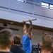A muscular man wearing a blue t-shirt is performing a dead hang or pull-up on a horizontal bar in what appears to be an indoor gymnasium or fitness facility. He has visible tattoos on his right arm and is wearing a watch on his left wrist. Several observers wearing dark clothing are watching from the foreground, with at least one wearing a patch-bearing uniform shirt.