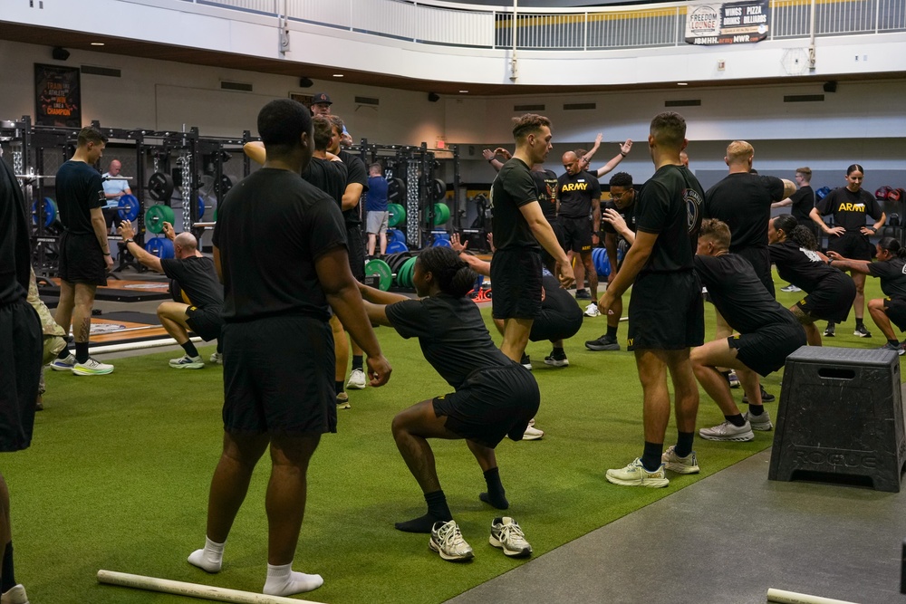 A large group of U.S. Army personnel are conducting a group physical training session inside a modern military fitness facility. Participants are uniformly dressed in black Army PT (Physical Training) uniforms and are performing squat exercises in synchronized rows. The facility appears well-equipped with weight racks, barbells, and turf flooring.
