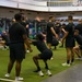 A large group of U.S. Army personnel are conducting a group physical training session inside a modern military fitness facility. Participants are uniformly dressed in black Army PT (Physical Training) uniforms and are performing squat exercises in synchronized rows. The facility appears well-equipped with weight racks, barbells, and turf flooring.