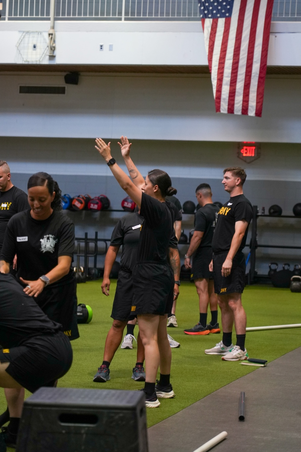 A group of individuals wearing matching black U.S. Army physical training uniforms are participating in a fitness session inside a military gymnasium. One person in the center is demonstrating an exercise with arms raised overhead. An American flag is prominently displayed on the upper wall of the facility.