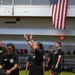 A group of individuals wearing matching black U.S. Army physical training uniforms are participating in a fitness session inside a military gymnasium. One person in the center is demonstrating an exercise with arms raised overhead. An American flag is prominently displayed on the upper wall of the facility.