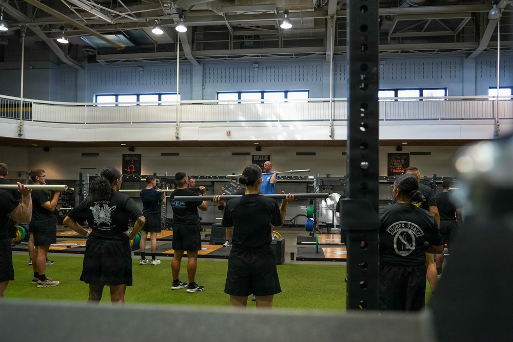 A group training session is taking place inside a large military or institutional gymnasium. An instructor in a blue shirt is demonstrating a barbell exercise to a group of participants dressed in matching black athletic uniforms. The facility features a two-level interior with an upper mezzanine track and professional weightlifting platforms.