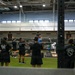 A group training session is taking place inside a large military or institutional gymnasium. An instructor in a blue shirt is demonstrating a barbell exercise to a group of participants dressed in matching black athletic uniforms. The facility features a two-level interior with an upper mezzanine track and professional weightlifting platforms.