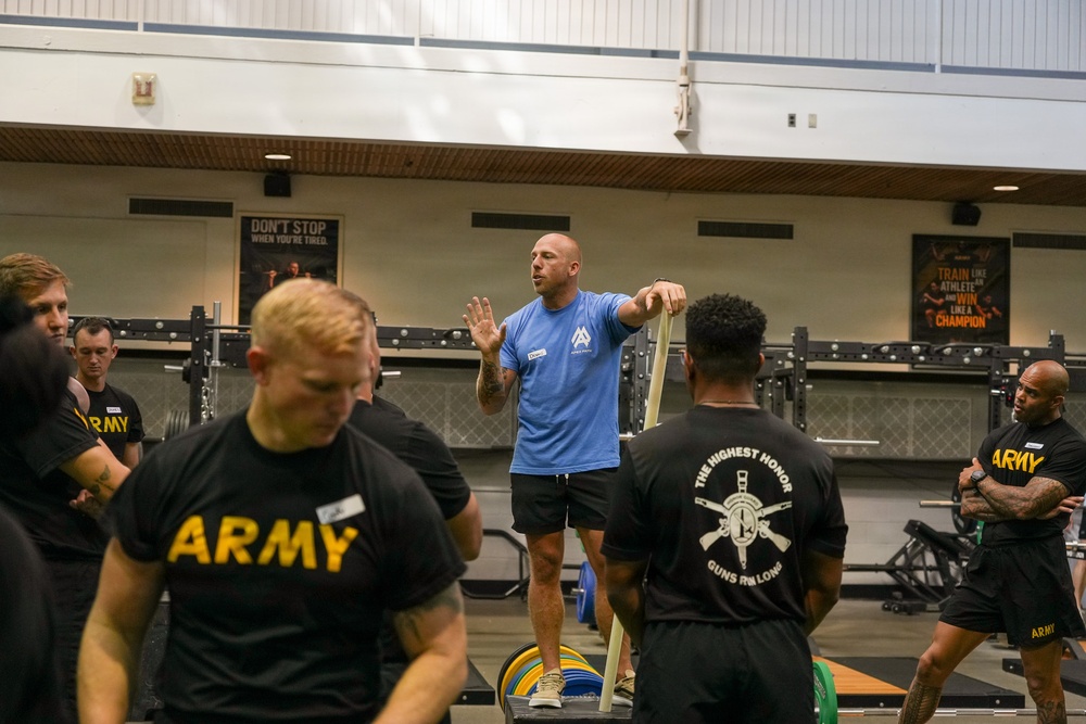A fitness instructor in a blue shirt is demonstrating resistance band techniques to a group of U.S. Army soldiers in a military gymnasium. The soldiers are wearing standard black Army PT (Physical Training) shirts and shorts. The setting appears to be a formal training session or clinic led by a civilian or contracted fitness professional.
