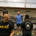 A fitness instructor in a blue shirt is demonstrating resistance band techniques to a group of U.S. Army soldiers in a military gymnasium. The soldiers are wearing standard black Army PT (Physical Training) shirts and shorts. The setting appears to be a formal training session or clinic led by a civilian or contracted fitness professional.