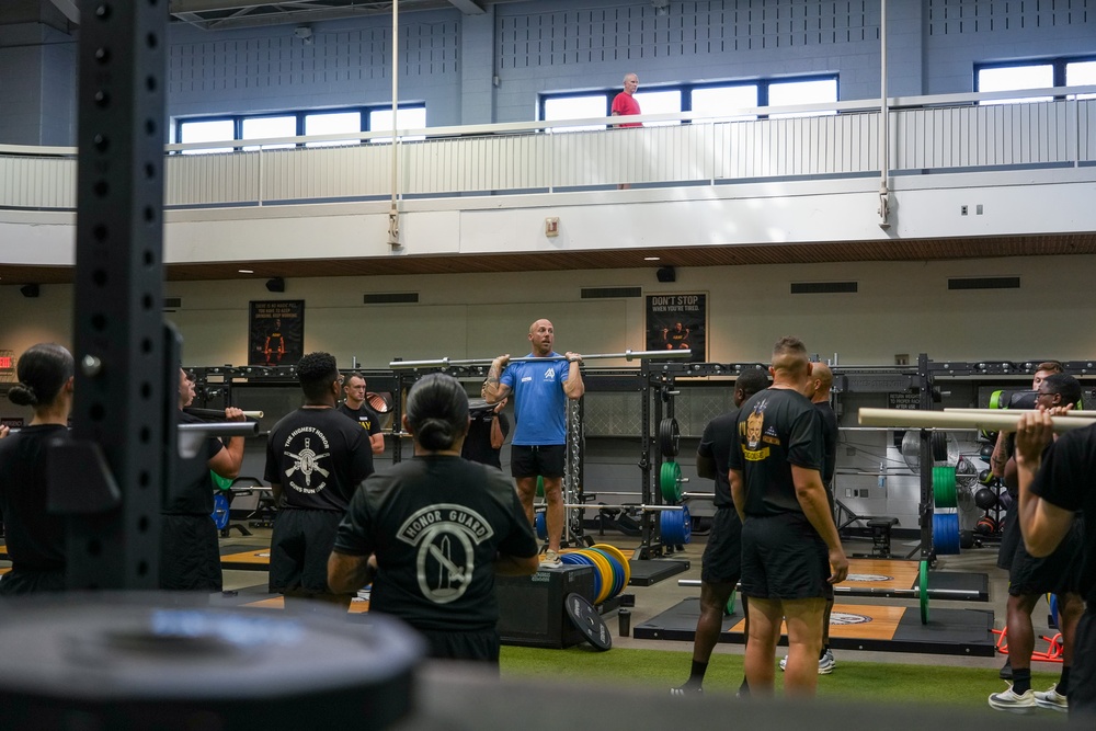 A group of military personnel in black PT uniforms are participating in a weightlifting training session inside a large gymnasium facility. A civilian instructor in a blue shirt is demonstrating a barbell exercise at the center of the group. The facility appears to be a well-equipped military fitness center with an upper observation level.