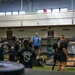 A group of military personnel in black PT uniforms are participating in a weightlifting training session inside a large gymnasium facility. A civilian instructor in a blue shirt is demonstrating a barbell exercise at the center of the group. The facility appears to be a well-equipped military fitness center with an upper observation level.