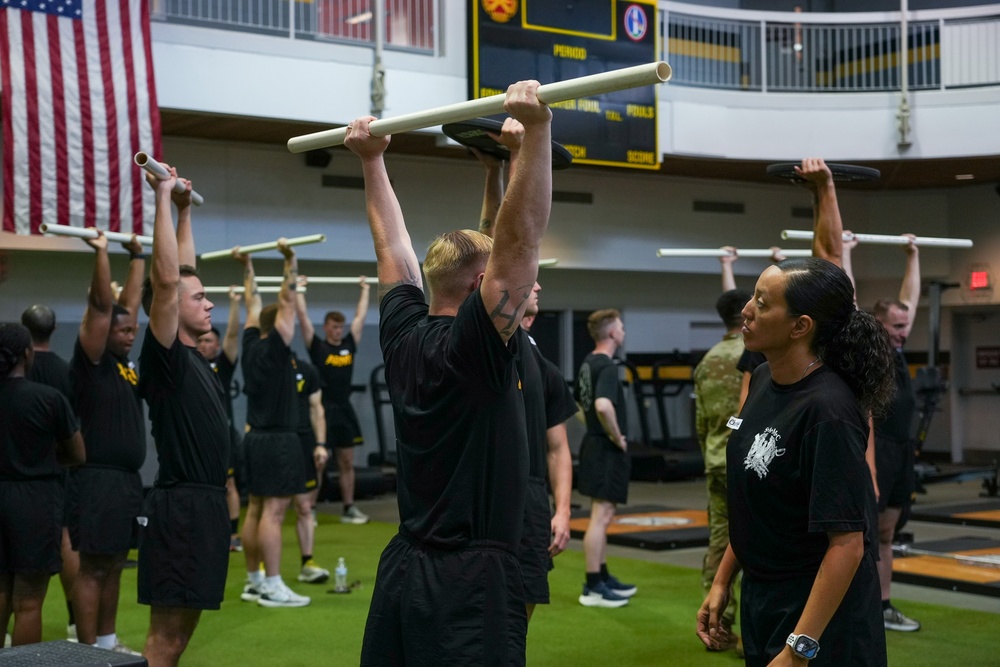 A large group of individuals in matching black athletic uniforms are participating in an organized fitness training session inside a military gymnasium. Participants are performing overhead press or lift exercises using white PVC pipes or light bars. An instructor or supervisor, a woman in a black t-shirt, is observing and monitoring the group.