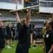 A large group of individuals in matching black athletic uniforms are participating in an organized fitness training session inside a military gymnasium. Participants are performing overhead press or lift exercises using white PVC pipes or light bars. An instructor or supervisor, a woman in a black t-shirt, is observing and monitoring the group.