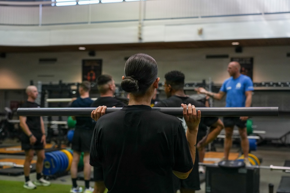 A group training session is taking place in a large indoor weight room or gymnasium. Multiple individuals wearing black athletic clothing are holding barbells across their shoulders, appearing to receive instruction from a coach or trainer dressed in a blue shirt. The foreground subject, viewed from behind, has hair in a bun and is holding an unloaded barbell in a front rack or back squat position.