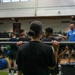 A group training session is taking place in a large indoor weight room or gymnasium. Multiple individuals wearing black athletic clothing are holding barbells across their shoulders, appearing to receive instruction from a coach or trainer dressed in a blue shirt. The foreground subject, viewed from behind, has hair in a bun and is holding an unloaded barbell in a front rack or back squat position.