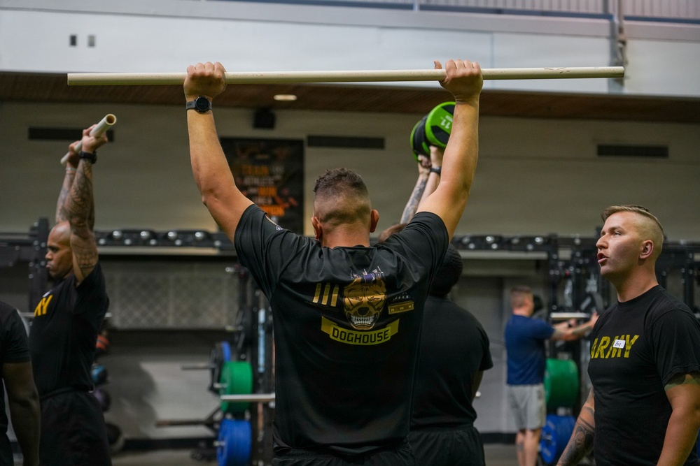 A group of individuals in a gym setting are engaged in a fitness training session. The central figure, wearing a black 'DOGHOUSE' t-shirt, is performing an overhead lift with a barbell or PVC pipe. Several participants are wearing U.S. Army-branded athletic shirts, suggesting this is a military physical training session.