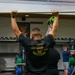 A group of individuals in a gym setting are engaged in a fitness training session. The central figure, wearing a black 'DOGHOUSE' t-shirt, is performing an overhead lift with a barbell or PVC pipe. Several participants are wearing U.S. Army-branded athletic shirts, suggesting this is a military physical training session.