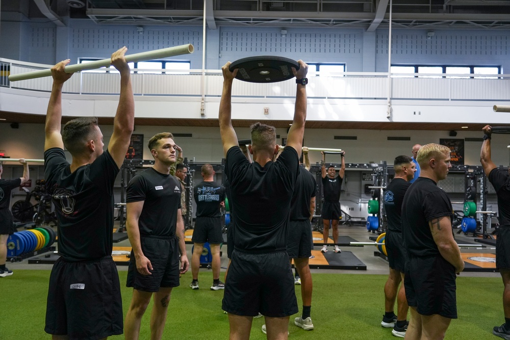 A group of individuals dressed in black athletic uniforms are participating in a structured strength and conditioning training session inside a large, well-equipped gymnasium. Several participants are performing overhead lifts using barbells, weight plates, and PVC pipes, while others observe or await their turn. The setting appears to be a military or institutional fitness facility based on the uniforms and organized group format.