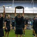 A group of individuals dressed in black athletic uniforms are participating in a structured strength and conditioning training session inside a large, well-equipped gymnasium. Several participants are performing overhead lifts using barbells, weight plates, and PVC pipes, while others observe or await their turn. The setting appears to be a military or institutional fitness facility based on the uniforms and organized group format.