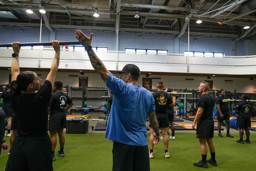 A group training session is taking place inside a large military fitness facility. A tattooed instructor in a blue shirt and black cap is demonstrating an overhead movement to a group of individuals wearing matching black military physical training uniforms. The facility is well-equipped with barbells, squat racks, and turf flooring.