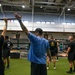 A group training session is taking place inside a large military fitness facility. A tattooed instructor in a blue shirt and black cap is demonstrating an overhead movement to a group of individuals wearing matching black military physical training uniforms. The facility is well-equipped with barbells, squat racks, and turf flooring.