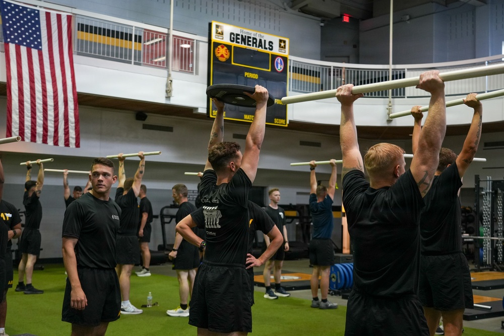 A group of individuals in matching black athletic uniforms are conducting a physical training session inside a military gymnasium. The facility displays 'Home of the Generals' on a scoreboard along with U.S. Army insignia and an American flag. Participants are performing various exercises including overhead bar hangs and weight plate lifts.