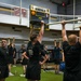 A group of individuals in matching black athletic uniforms are conducting a physical training session inside a military gymnasium. The facility displays 'Home of the Generals' on a scoreboard along with U.S. Army insignia and an American flag. Participants are performing various exercises including overhead bar hangs and weight plate lifts.
