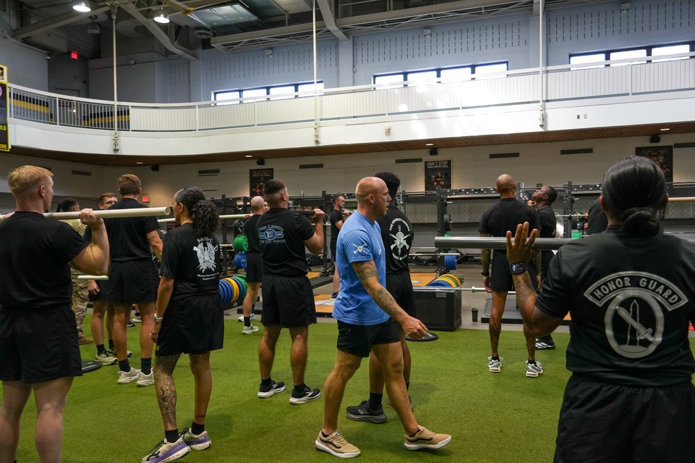 A group of military personnel are engaged in a physical training session inside a large gymnasium facility. Participants are performing barbell exercises, with several individuals holding barbells across their shoulders. One person wearing a blue shirt appears to be an instructor or trainer directing the group.