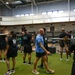 A group of military personnel are engaged in a physical training session inside a large gymnasium facility. Participants are performing barbell exercises, with several individuals holding barbells across their shoulders. One person wearing a blue shirt appears to be an instructor or trainer directing the group.