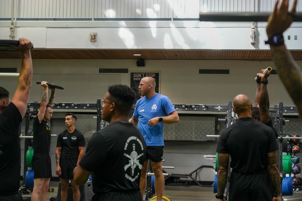 A fitness training session is taking place in a well-equipped gymnasium. A man in a blue t-shirt appears to be instructing or coaching a group of individuals dressed predominantly in black athletic wear. Several participants are performing overhead exercises with various implements including barbells and what appear to be maces or clubs.