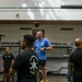 A fitness training session is taking place in a well-equipped gymnasium. A man in a blue t-shirt appears to be instructing or coaching a group of individuals dressed predominantly in black athletic wear. Several participants are performing overhead exercises with various implements including barbells and what appear to be maces or clubs.