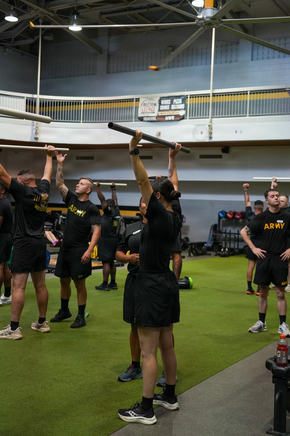 A group of U.S. Army soldiers in standard black PT (Physical Training) uniforms are conducting an indoor fitness training session inside what appears to be a military fitness facility. Several soldiers are holding bars or poles overhead as part of a structured exercise drill. A banner in the background references 'Freedom Lounge' and 'Army MWR,' indicating this is a military installation recreational facility.