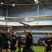 A group of U.S. Army soldiers in standard black PT (Physical Training) uniforms are conducting an indoor fitness training session inside what appears to be a military fitness facility. Several soldiers are holding bars or poles overhead as part of a structured exercise drill. A banner in the background references 'Freedom Lounge' and 'Army MWR,' indicating this is a military installation recreational facility.