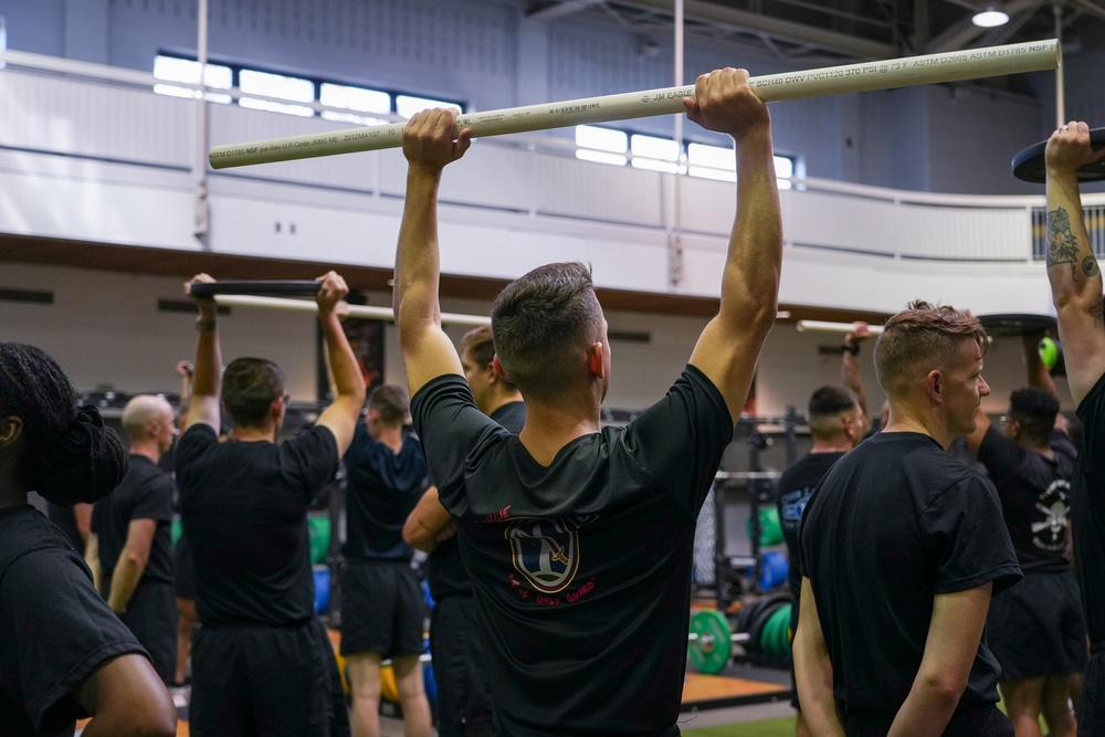 A large group of individuals dressed in black athletic wear are participating in a group fitness or physical training session inside a gymnasium. The central figure is holding a PVC pipe overhead, likely performing an overhead press or mobility drill. The setting appears to be a military or organized athletic training facility based on the uniformity of attire and structured group format.