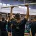 A large group of individuals dressed in black athletic wear are participating in a group fitness or physical training session inside a gymnasium. The central figure is holding a PVC pipe overhead, likely performing an overhead press or mobility drill. The setting appears to be a military or organized athletic training facility based on the uniformity of attire and structured group format.