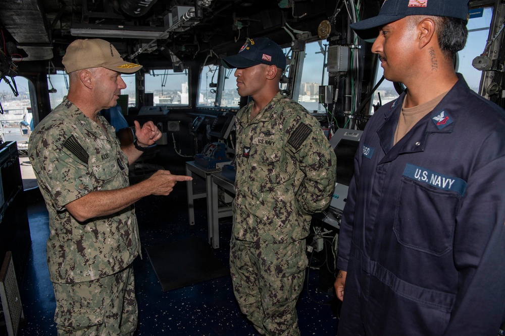 Three U.S. Navy personnel are engaged in conversation aboard what appears to be the bridge of a naval vessel. A senior officer in NWU Type III camouflage uniform with a gold-brimmed cap is speaking to two junior sailors, one in camouflage utilities and one in navy blue coveralls. The setting appears to be an active ship's bridge with navigation equipment visible in the background.