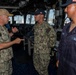 Three U.S. Navy personnel are engaged in conversation aboard what appears to be the bridge of a naval vessel. A senior officer in NWU Type III camouflage uniform with a gold-brimmed cap is speaking to two junior sailors, one in camouflage utilities and one in navy blue coveralls. The setting appears to be an active ship's bridge with navigation equipment visible in the background.