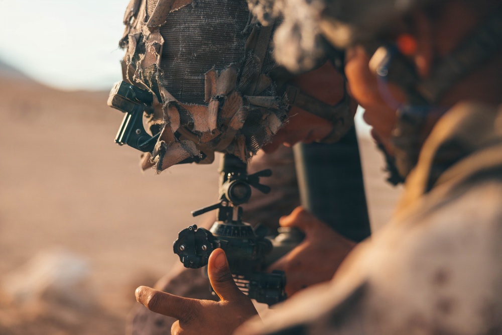A soldier in desert camouflage gear is closely examining or adjusting a weapon-mounted optical scope or sight system. The individual is wearing a heavily camouflaged helmet with netting and attached accessories, consistent with modern military field equipment. The arid, sandy background suggests a desert operational environment.