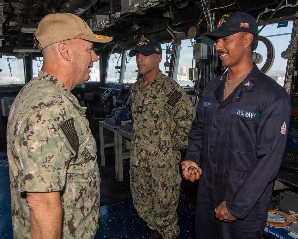 Three U.S. Navy personnel are engaged in conversation aboard what appears to be the bridge of a naval vessel. Two sailors are in Navy Working Uniform (NWU) camouflage pattern while one is in navy blue coveralls. The setting is clearly a ship's bridge with navigation equipment and windows visible in the background.