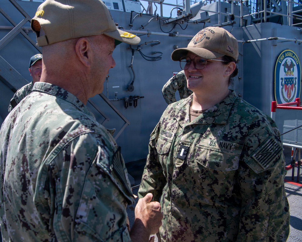 Two U.S. Navy personnel in NWU Type III (woodland camouflage) uniforms are engaged in conversation or a handshake aboard what appears to be the USS Ashland (LSD-48), a dock landing ship. The female sailor on the right displays a rank insignia consistent with an Ensign or Lieutenant Junior Grade, and both wear tan baseball-style covers. A third service member is partially visible in the background.