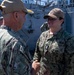 Two U.S. Navy personnel in NWU Type III (woodland camouflage) uniforms are engaged in conversation or a handshake aboard what appears to be the USS Ashland (LSD-48), a dock landing ship. The female sailor on the right displays a rank insignia consistent with an Ensign or Lieutenant Junior Grade, and both wear tan baseball-style covers. A third service member is partially visible in the background.