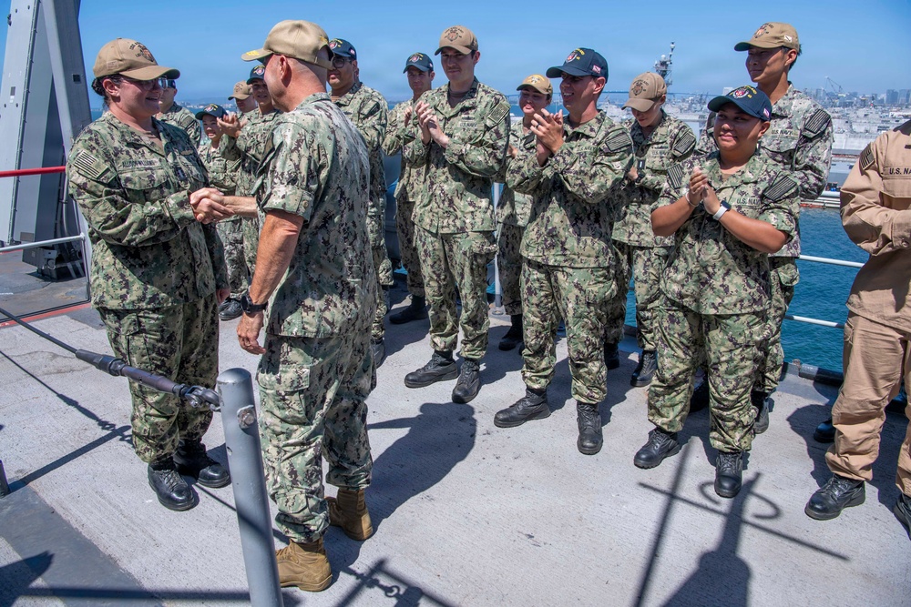 A group of U.S. Navy personnel in NWU Type III (woodland camouflage) uniforms are gathered on the deck of a naval vessel, applauding a fellow service member who appears to be receiving recognition or a commendation. The ceremony is taking place outdoors on a ship deck with a harbor and cityscape visible in the background. One individual on the far right is wearing tan/desert camouflage, distinguishing them from the rest of the group.