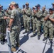 A group of U.S. Navy personnel in NWU Type III (woodland camouflage) uniforms are gathered on the deck of a naval vessel, applauding a fellow service member who appears to be receiving recognition or a commendation. The ceremony is taking place outdoors on a ship deck with a harbor and cityscape visible in the background. One individual on the far right is wearing tan/desert camouflage, distinguishing them from the rest of the group.
